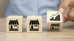 Blocos de madeira com ícones de lojas alinhados sobre uma mesa; uma mão gira o último bloco, que mostra um gráfico de crescimento, simbolizando expansão de negócios.